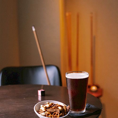 A glass of beer and a bowl of mixed nuts on a table, with pool cues and a chair in the background.