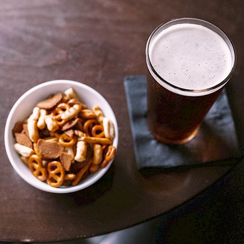 A bowl of mixed snacks and a pint of beer on a coaster are on a wooden table.