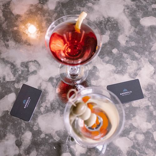 Two cocktails in martini glasses on a marble surface with two black cards beside them, featuring a lemon twist and olives garnishes.