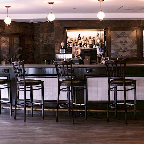 An empty bar area with high chairs, pendant lighting, and a well-stocked liquor shelf in the background. The flooring is wooden, and no people are visible.