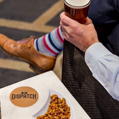 A person in dress clothes, sitting and holding a pint of beer. Colorful socks and dress shoes are visible. A bowl of nuts and candle are on the table.