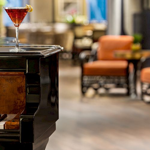 A cocktail glass sits on a grand piano in a cozy lounge area with orange cushioned chairs and soft focus background.