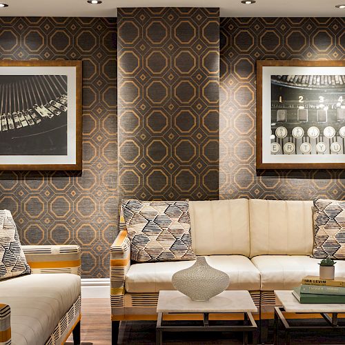 A modern living room with geometric wallpaper, white couches, patterned cushions, framed black-and-white photos, a vase, and stacked books on tables.